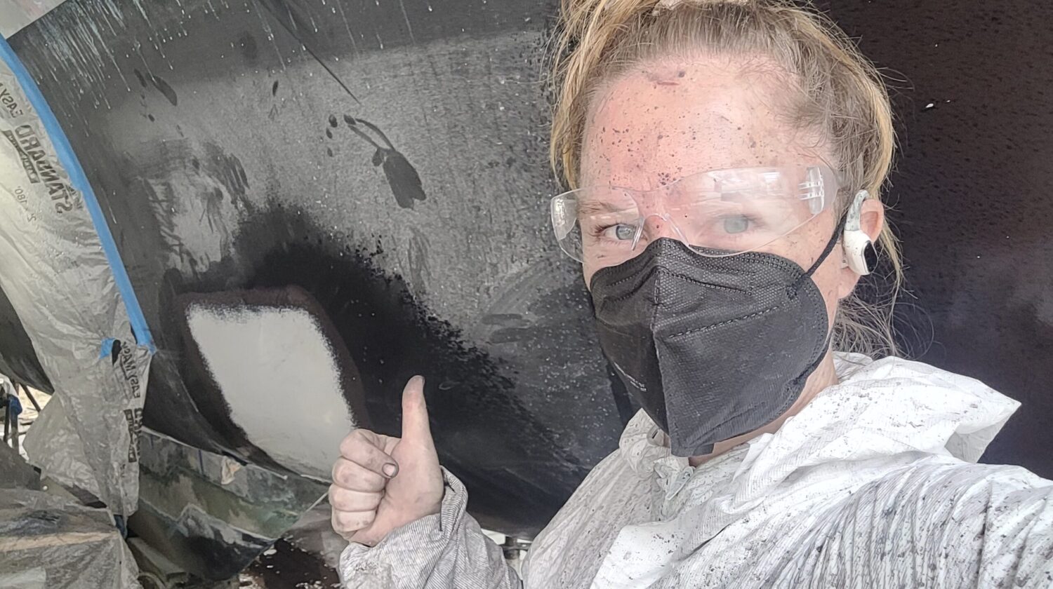 Woman wearing PPE gives thumbs up with antifoul on her face and a boat hull behind her