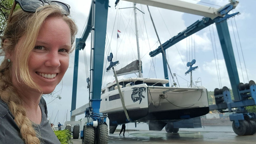 Smiling woman with sunglasses watches a yacht being lifted by a blue hoist at a marina. Sky is cloudy, and water is on the ground.