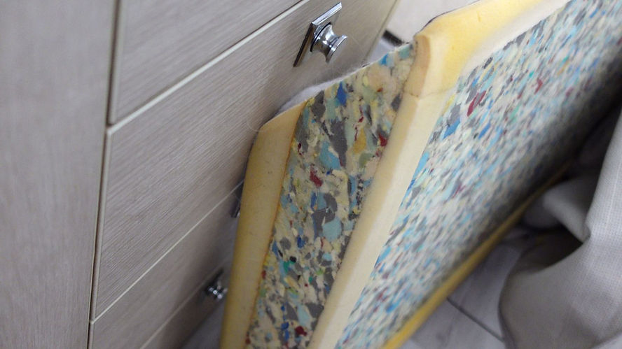 Foam mattress made of glued foam chips leaning against a wooden drawer unit.