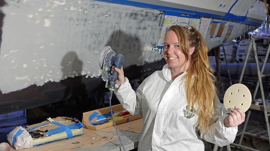 Woman in white coveralls smiling while holding a sander and disc, working on a boat hull in a workshop, background shows tools and ladders.