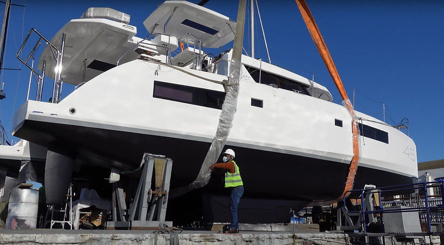 A boat is being hoisted with straps at a shipyard. A worker in a helmet and vest oversees. The sky is clear and blue.