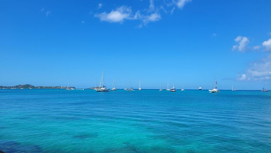 Sailboats float on clear blue ocean in St Martin under a bright blue sky with scattered clouds. A distant shoreline Anguilla is visible in the background.