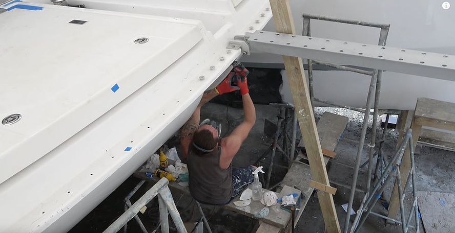 Person in a tank top works on a white boat deck with tools. Scaffolding, wooden beams, and scattered equipment set the scene.
