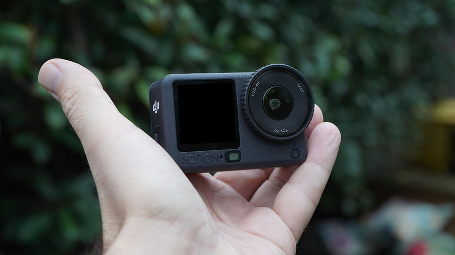 A hand holds a Osmo 6 action camera with a round lens. The background is blurred greenery.