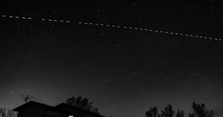 Night sky with stars and a dotted line of lights, possibly a satellite, over silhouettes of trees and a house with an antenna.