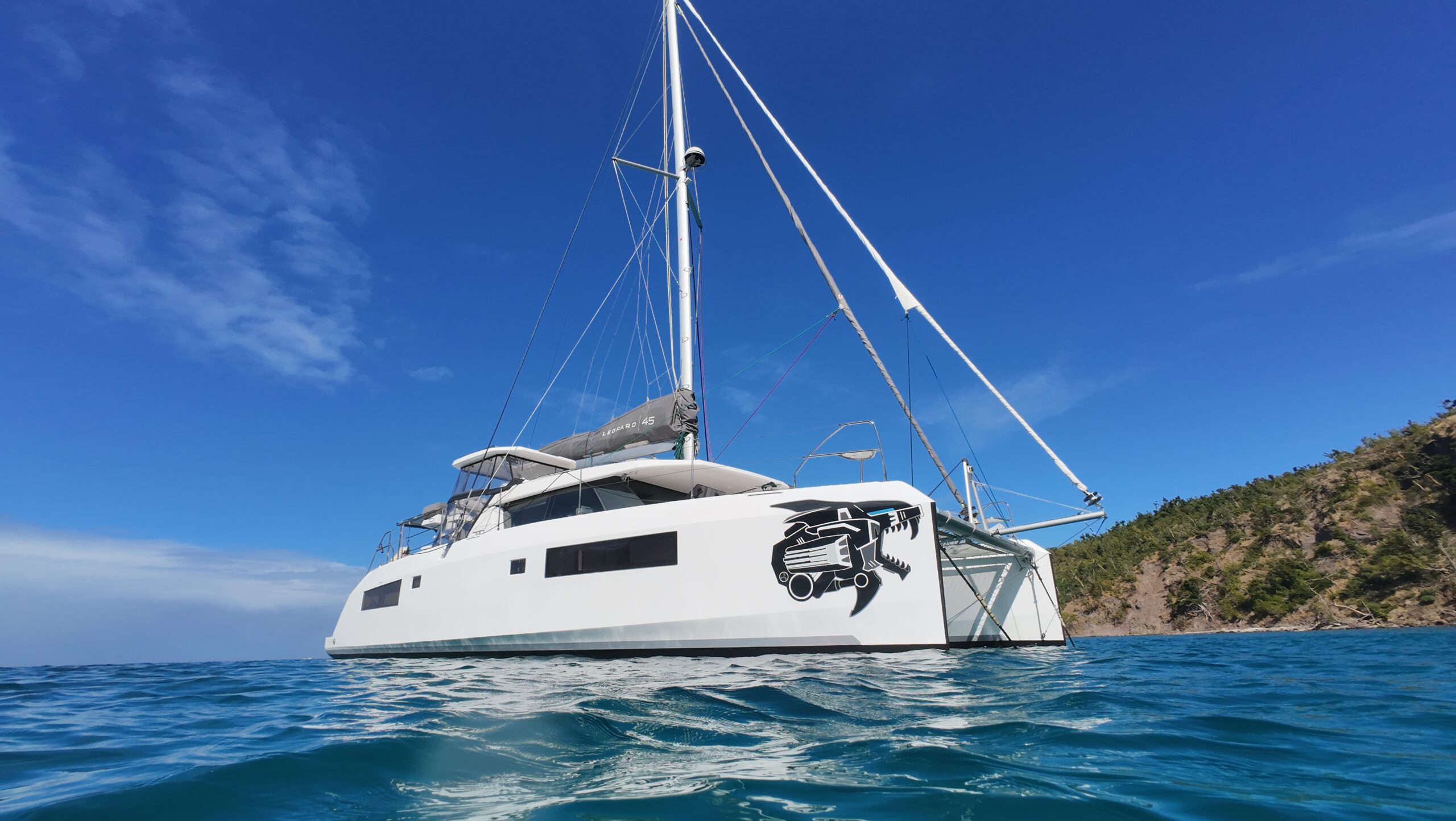 White Leopard 45 catamaran anchored in tropical bay with blue skies, blue water and lush greenery. The yacht has a decal on the front indicating it SV Liger from Travel Sketch
