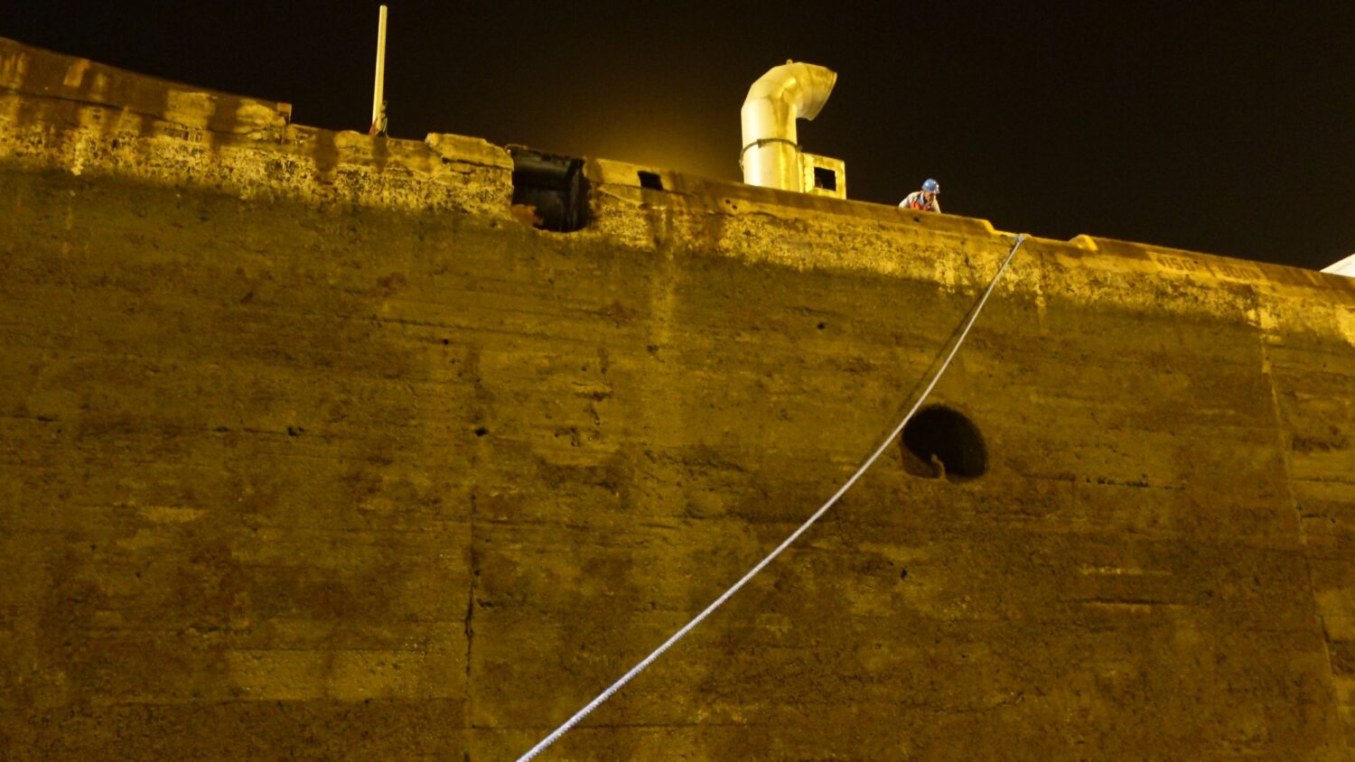 Rope going up the wall of the Panama Canal to a man at the top wearing a hard hat and high vis uniform